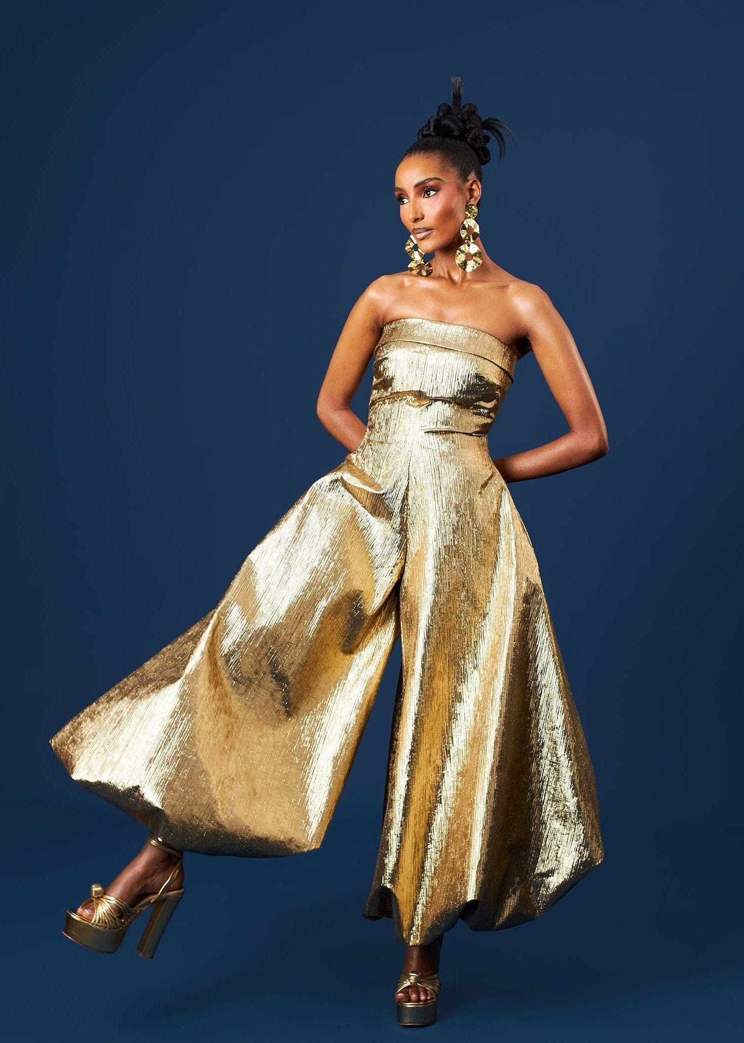 Woman wearing a gold strapless jumpsuit against a blue background